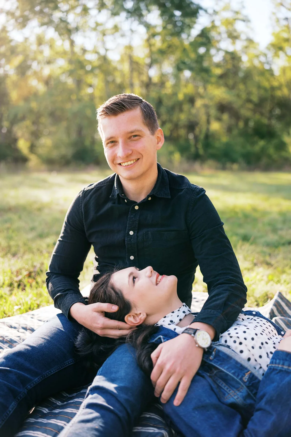 Mosolygó pár ül a fűben, egyikük a másik ölébe hajtja a fejét.