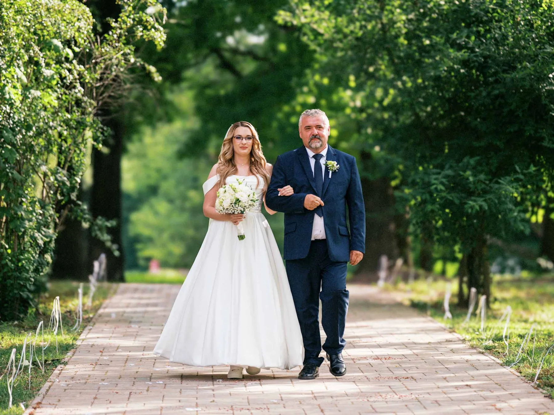 Menyasszony és vőlegény kézen fogva sétálnak egy fasoron.
